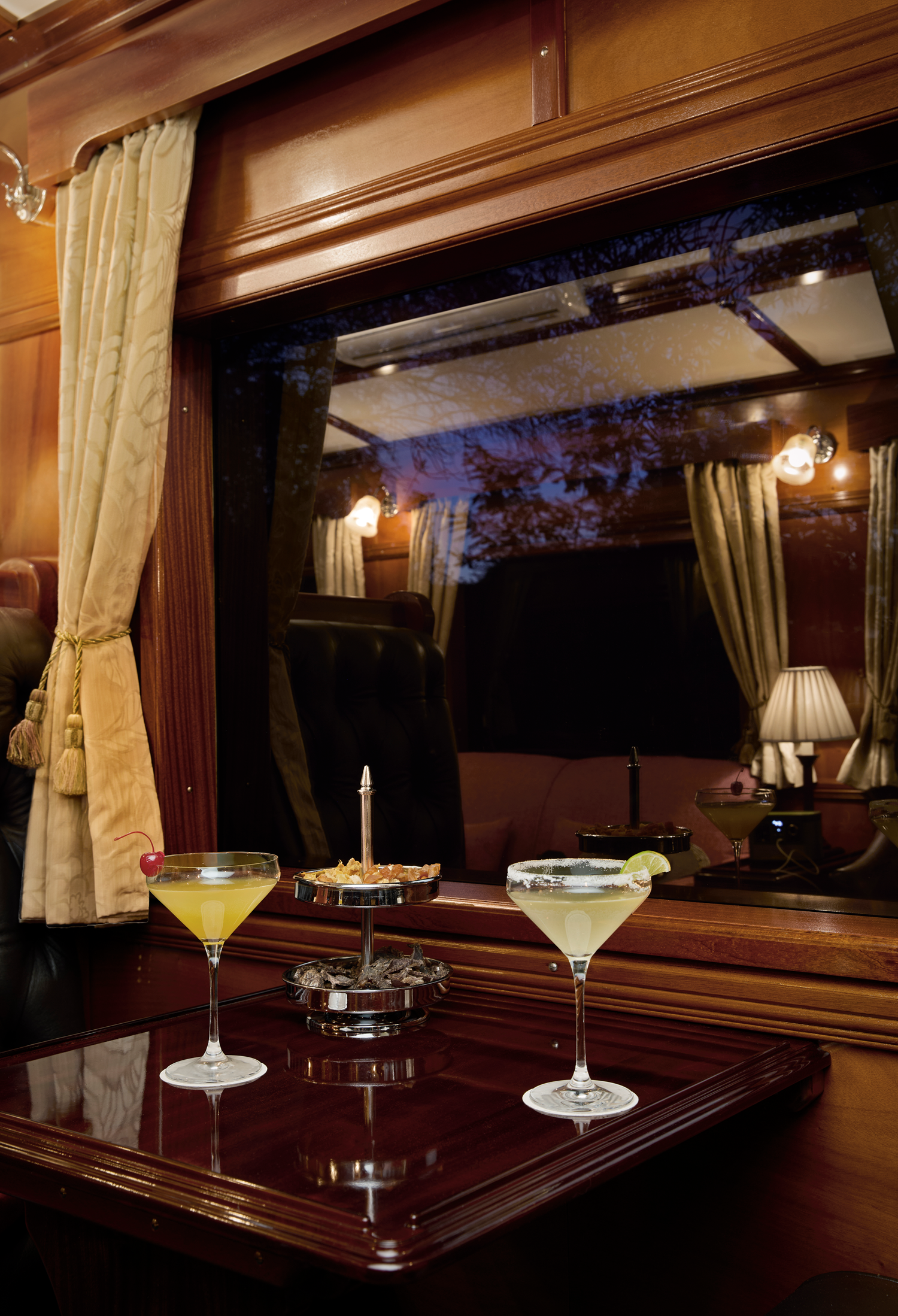 Cocktails auf einem Tisch im luxuriösen Rovos Rail mit Blick aus dem Zugfenster in die afrikanische Nacht.
