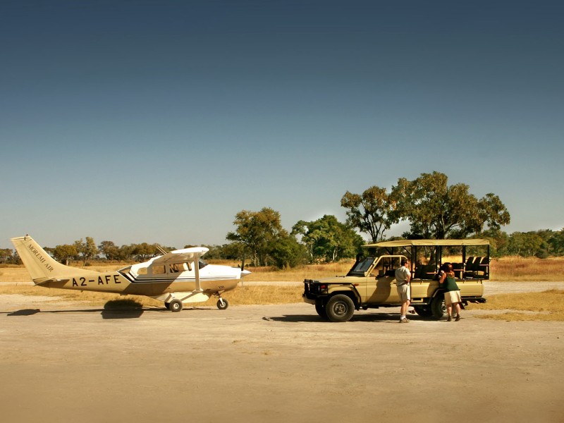 Kleines Buschflugzeug und Safari-Fahrzeug auf einer Landepiste in der Wildnis Botswanas. Reisende wechseln vom Flugzeug in den Geländewagen und beginnen ihre Flugsafari zu den abgelegenen Naturparadiesen des Okavango-Deltas und anderer Schutzgebiete.