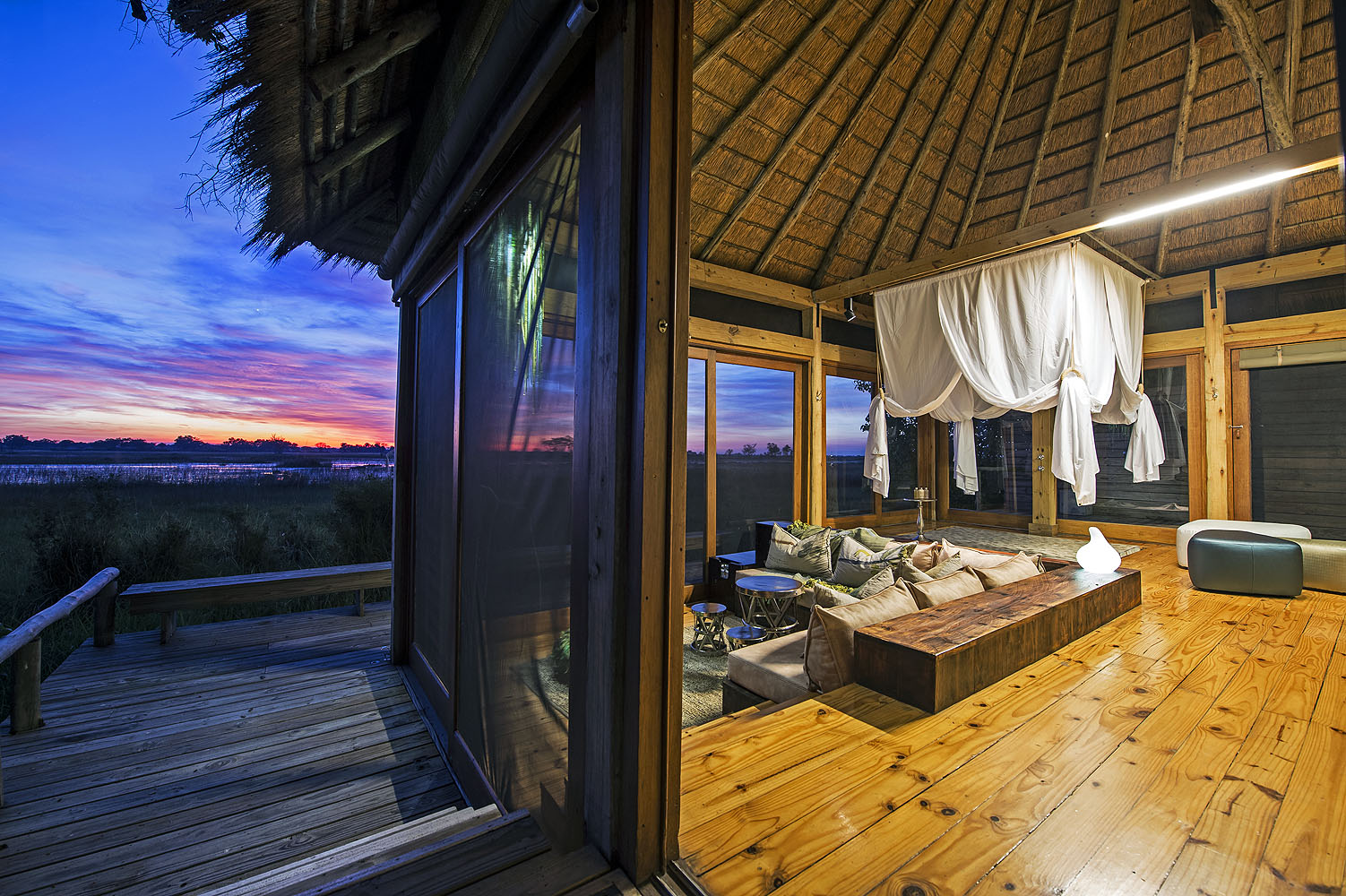 Stilvolle Safari-Lodge mit offenem Wohnbereich und Blick auf die Wasserlandschaft des Okavango-Deltas in Botswana während der Abenddämmerung.