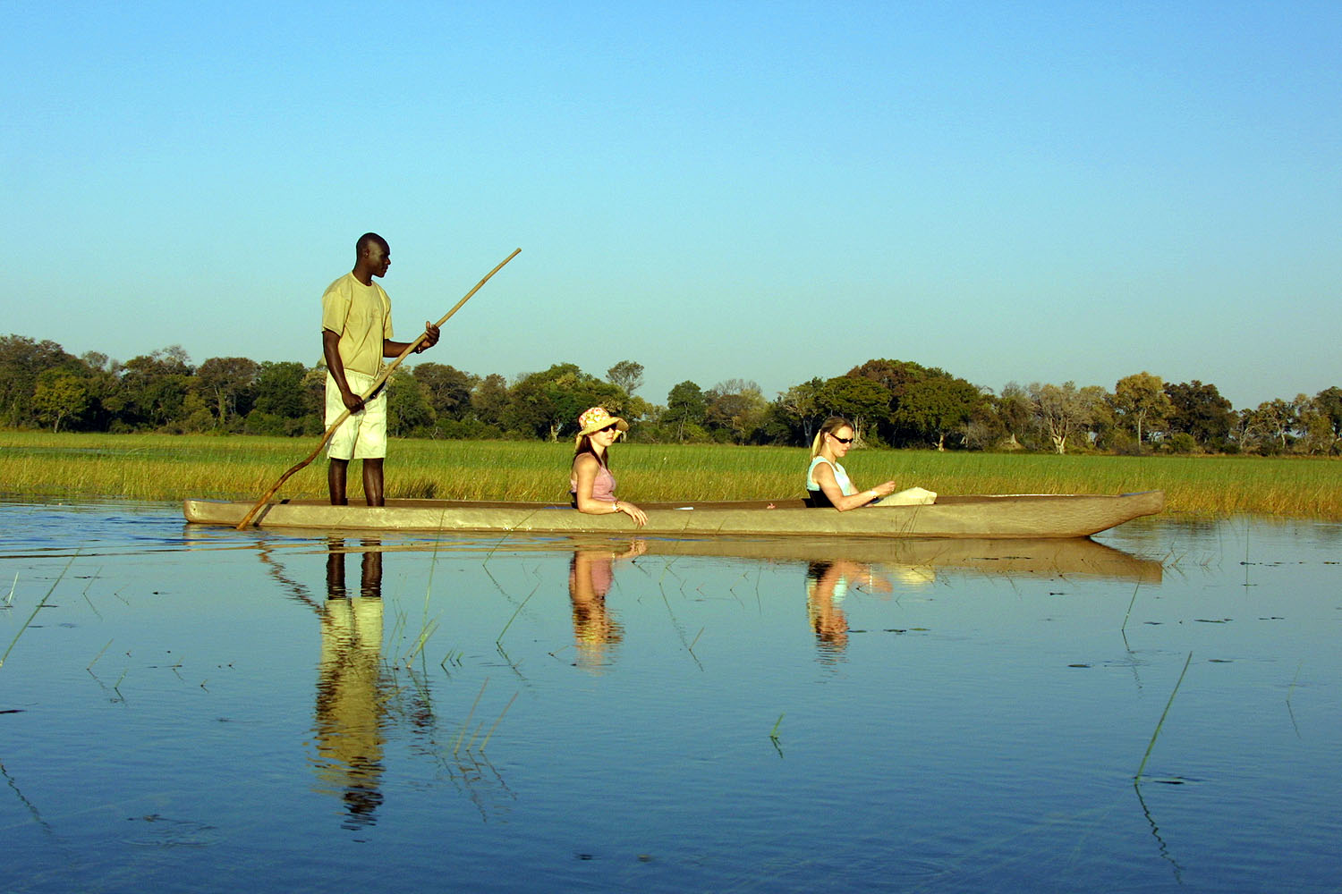 Reisende gleiten in einem traditionellen Mokoro-Einbaumboot durch die ruhigen Wasserwege des Okavango-Deltas in Botswana. Ein einheimischer Poler steuert das Boot durch die weite Schilflandschaft, die sich im klaren Wasser spiegelt.