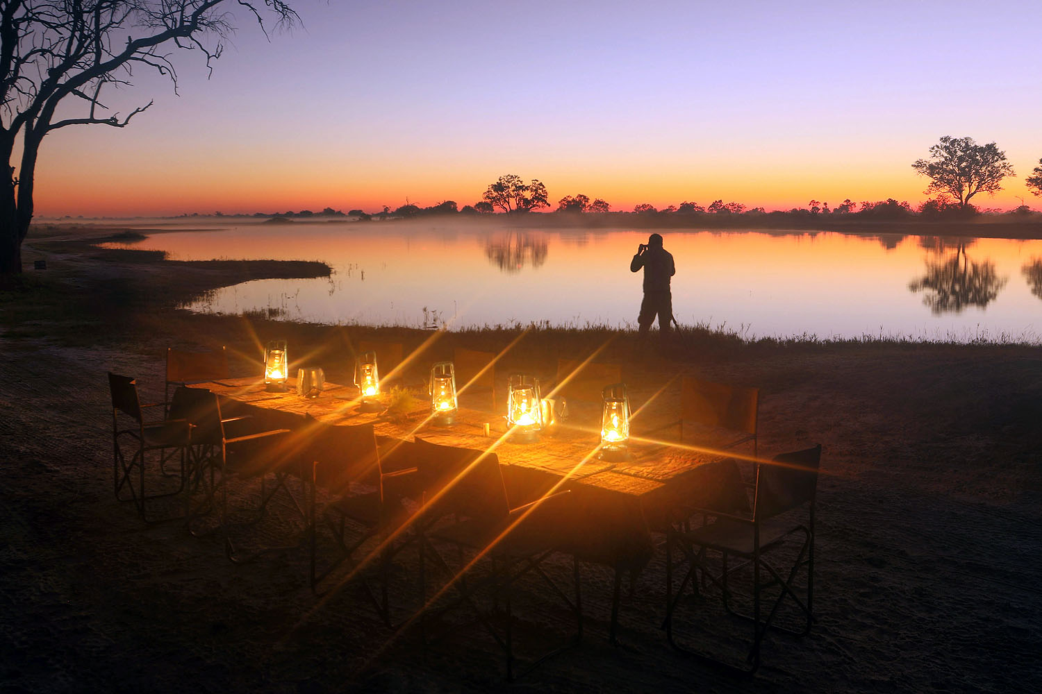 Romantisch gedeckter Tisch mit Laternen am Ufer eines Wasserlochs in Botswana während eines farbenprächtigen Sonnenuntergangs.