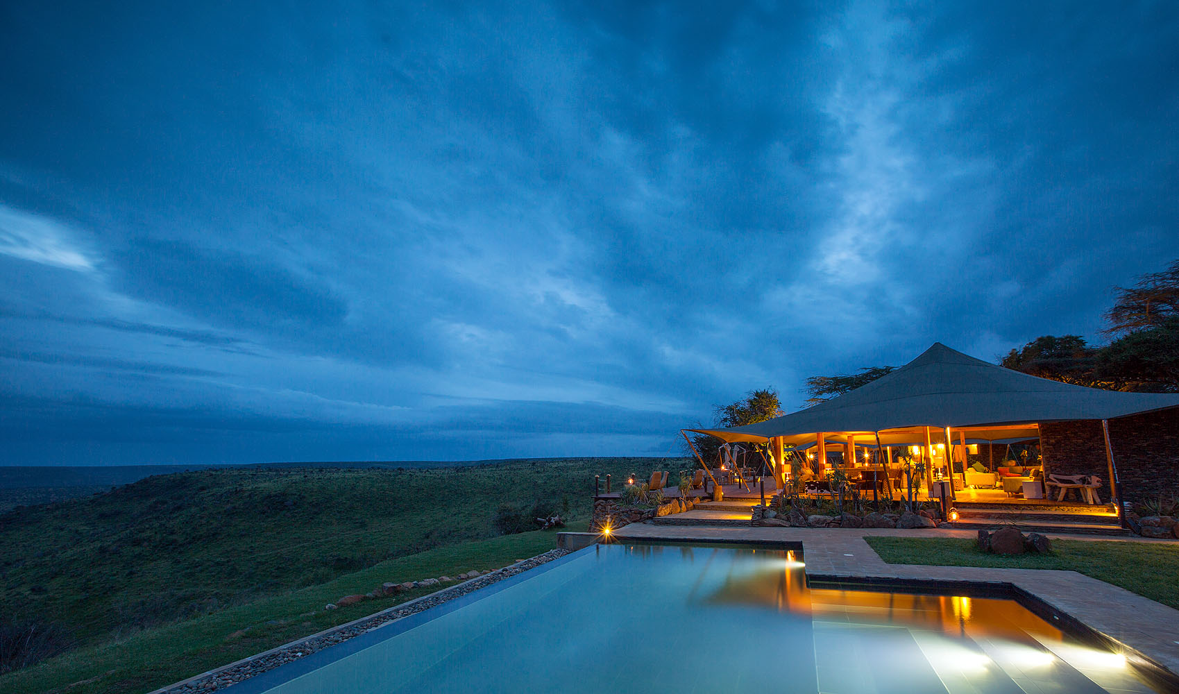 Exklusive Safari-Lodge mit Infinity-Pool auf einer Anhöhe und Panoramablick über die afrikanische Savanne bei Sonnenuntergang.