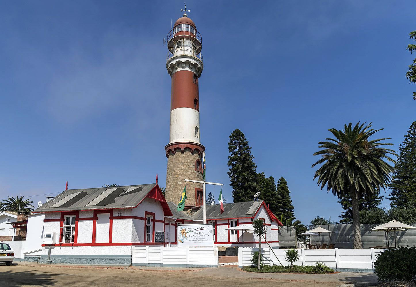 Historischer rot-weißer Leuchtturm von Swakopmund in Namibia mit Palmen und kolonialer Architektur unter blauem Himmel.