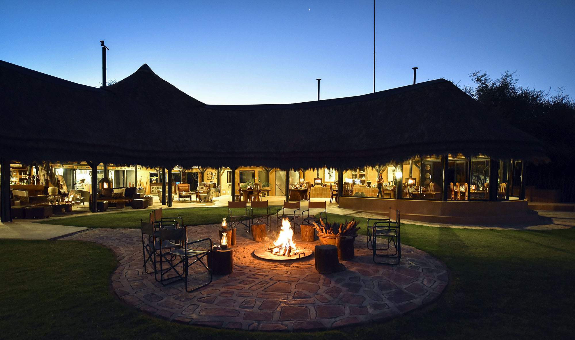 Stilvolle Safari-Lodge in Namibia bei Nacht mit beleuchtetem Hauptgebäude und gemütlicher Feuerstelle unter dem klaren Abendhimmel.