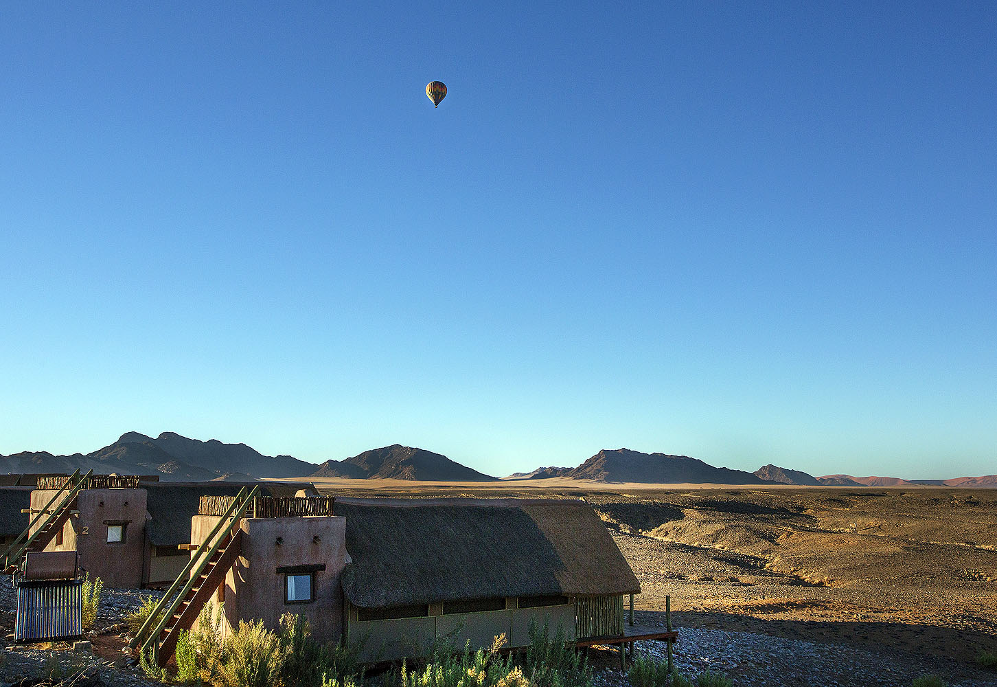 Komfortable Wüstenlodge in Namibia mit Blick auf karge Berglandschaften und einen Heißluftballon über der weiten Namib-Wüste.